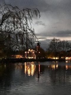 Evening lake with warm lights reflecting on the water beside trees and a building under cloudy skies.