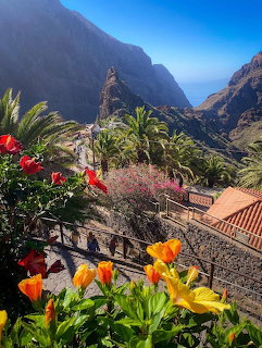 Colorful flowers frame mountain village nestled in dramatic gorge with palm trees and traditional buildings