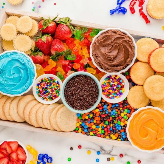 Overhead view of dessert board with cookies, strawberries, frosting bowls, and colorful sprinkles on white marble