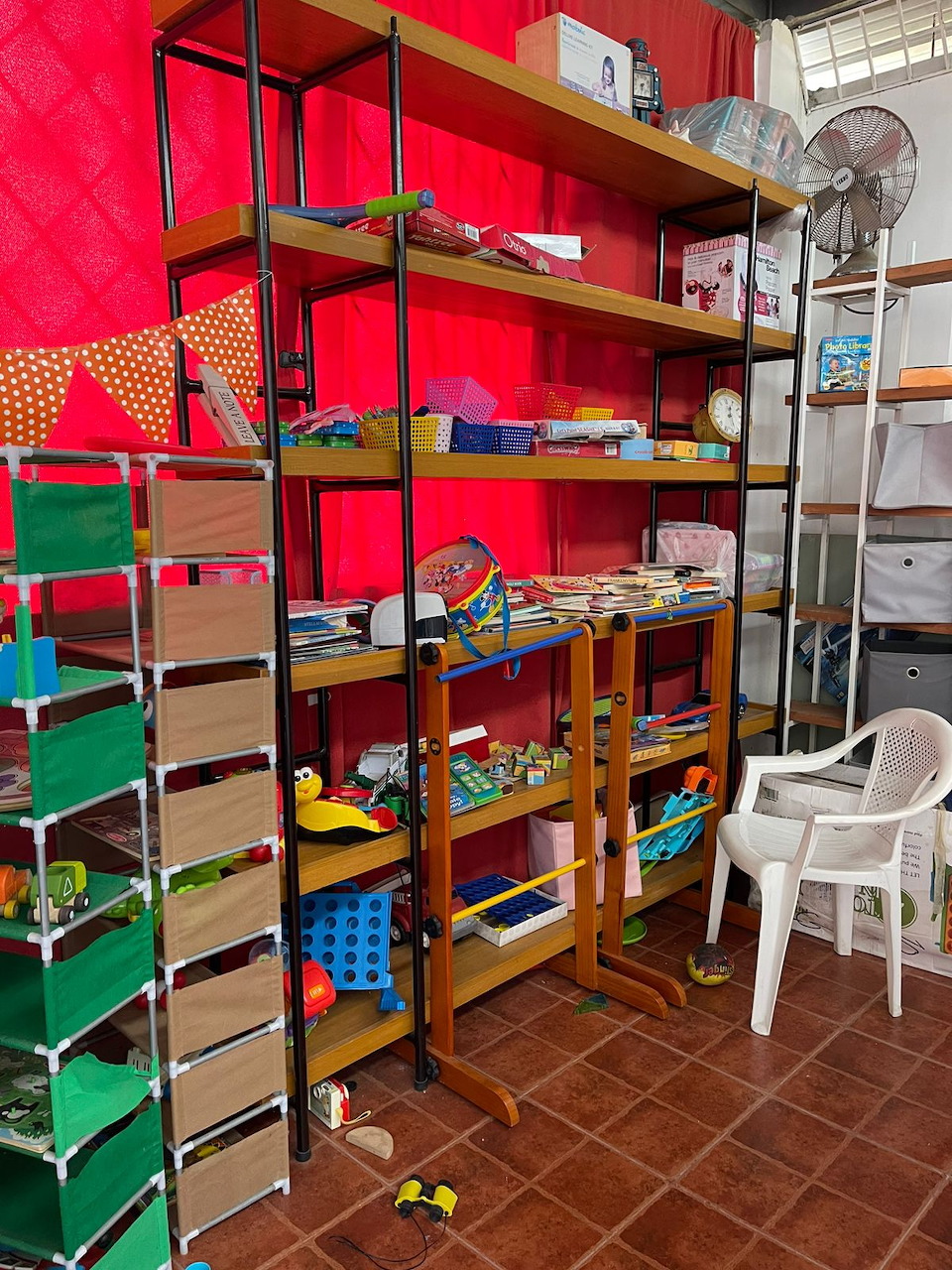 Wooden shelves with school supplies and books in a colorful classroom with a red wall and white chair.