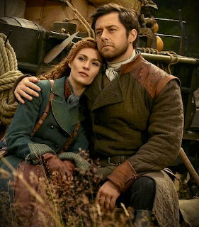 Couple in historical clothing sitting together outdoors near a wagon with ropes