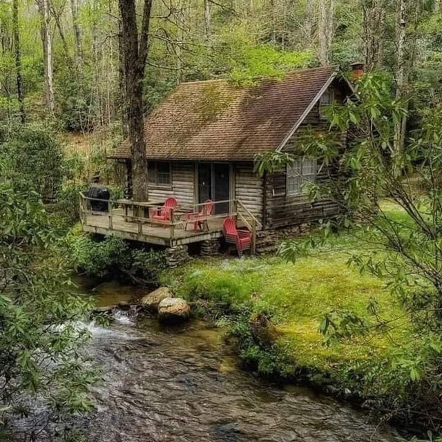 Rustic log cabin beside a creek in a wooded area with a deck and red chairs