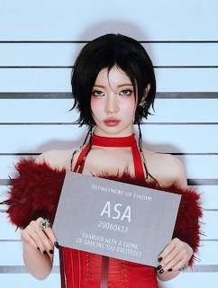 Woman in Asa cosplay with black hair and red outfit holding character identification sign