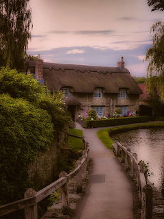 Thatched cottage beside stream with lush garden and pathway at dusk