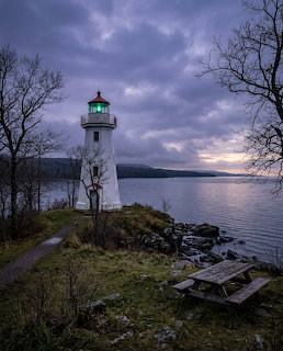 White lighthouse with green light on a rocky shoreline at dusk with calm water and overcast sky
