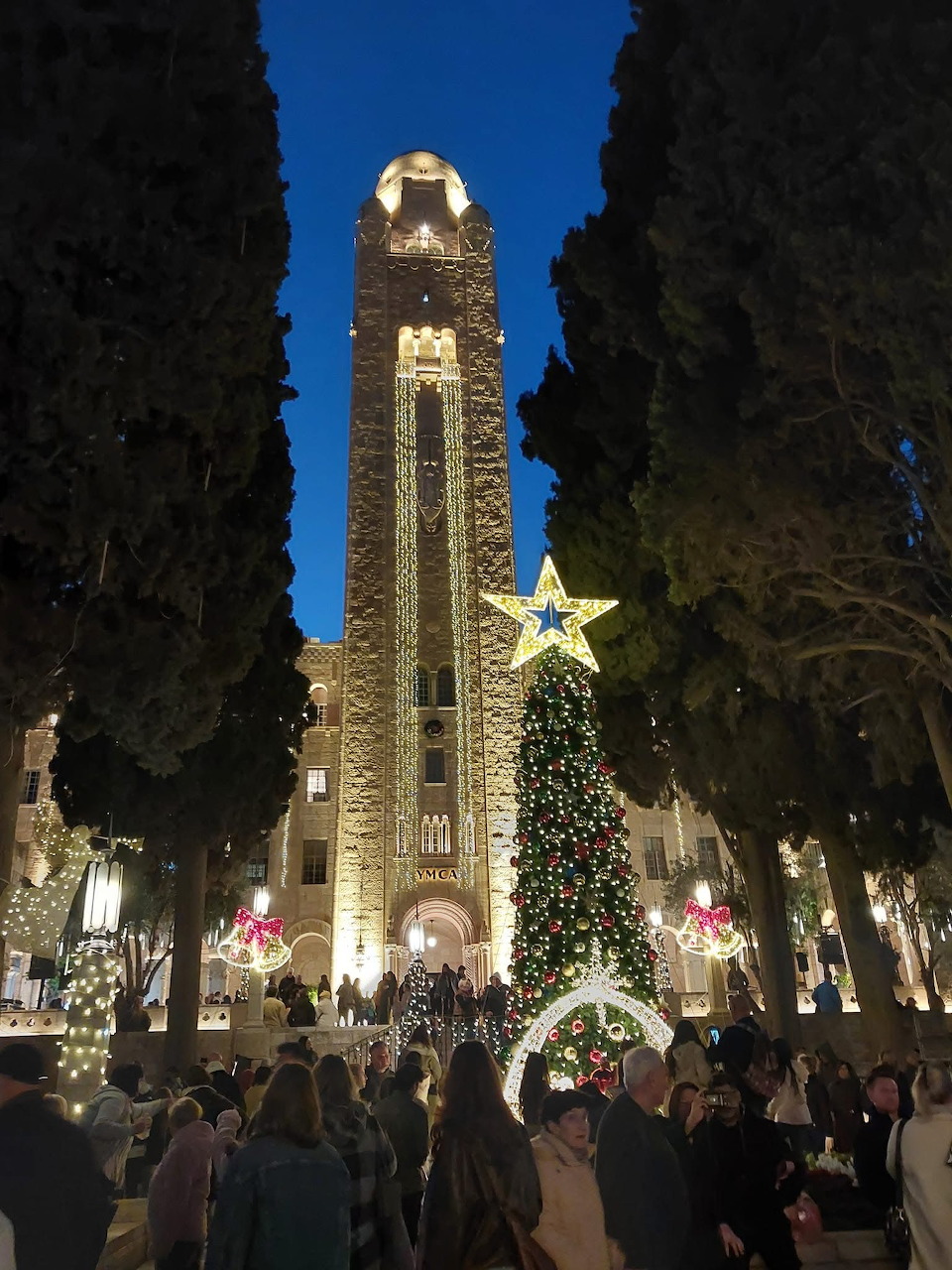 Illuminated bell tower and Christmas tree with star at dusk in Jerusalem, surrounded by holiday lights and people