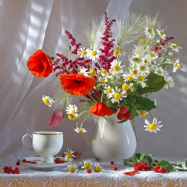 White pitcher with red poppies, daisies, and wildflowers next to a teacup on a table with white fabric