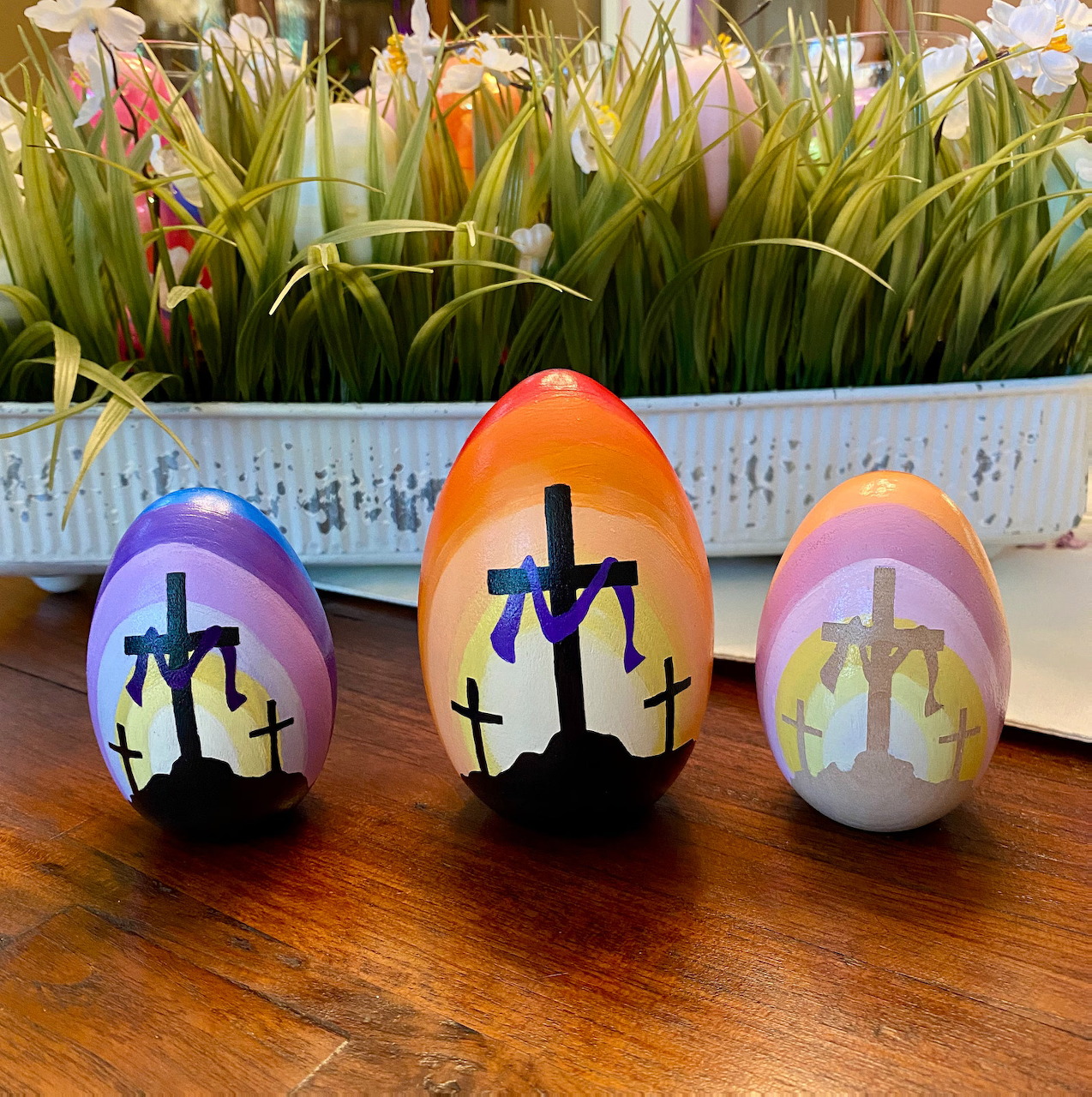 Three painted Easter eggs with cross silhouettes against colorful sunset skies, arranged on wooden table with flowers behind