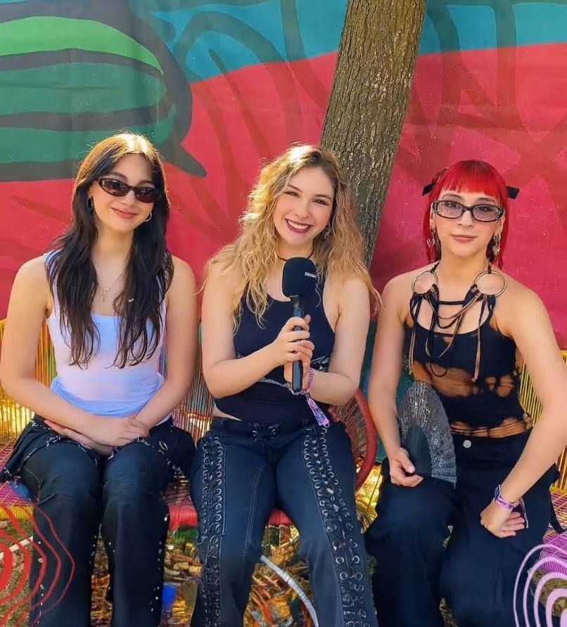 Three young women seated outdoors against a colorful mural, one holding a microphone