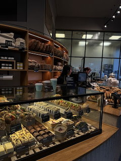 Coffee shop interior with pastry display case, wooden shelves of bread, and a barista at the counter