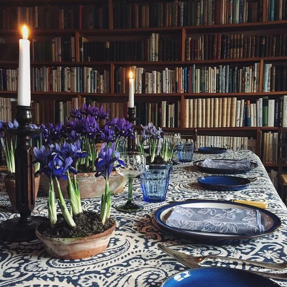 Formal dining table with blue dishes and purple flowers in front of wooden bookshelves
