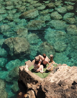 Two people having a picnic on rocky outcrop above clear turquoise ocean water