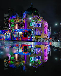 Flinders Street Station lit with colorful lights at night, mirrored in rain-soaked street