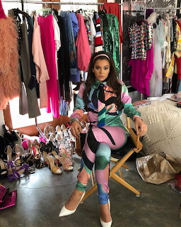 Woman in colorful geometric outfit sitting in director's chair in fashion showroom with clothing racks