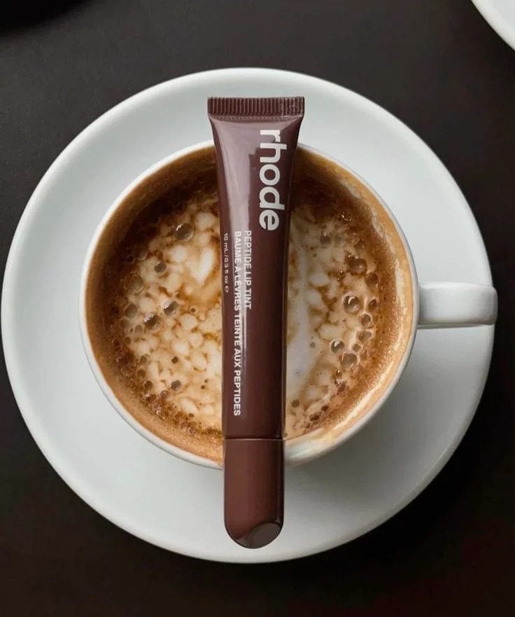 Brown Rhode lip gloss tube resting on white cup of cappuccino with foam