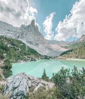 Turquoise alpine lake nestled in rocky Dolomite mountains with evergreen trees and dramatic peaks under cloudy blue sky