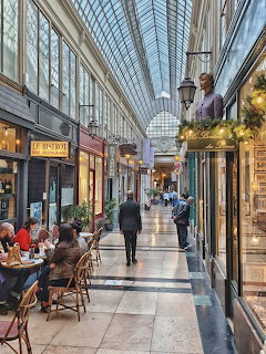 Glass-roofed shopping arcade with cafés, shops, and people in a long indoor passage