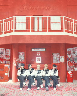 Six K-pop boys in matching outfits sitting on a bench in front of a red building with heart decorations