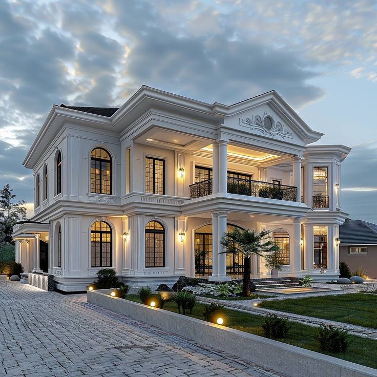 White two-story mansion with columns and arched windows lit up at dusk with landscaped grounds