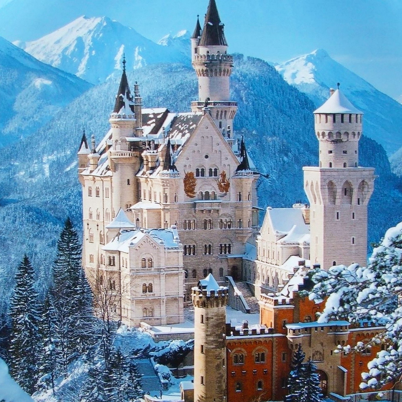 Snow-covered Neuschwanstein Castle with tall towers against blue mountain peaks and pine forest in winter