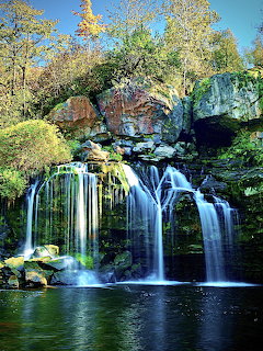 Waterfall flowing over mosy rocks into a dark pool surrounded by trees with autumn foliage