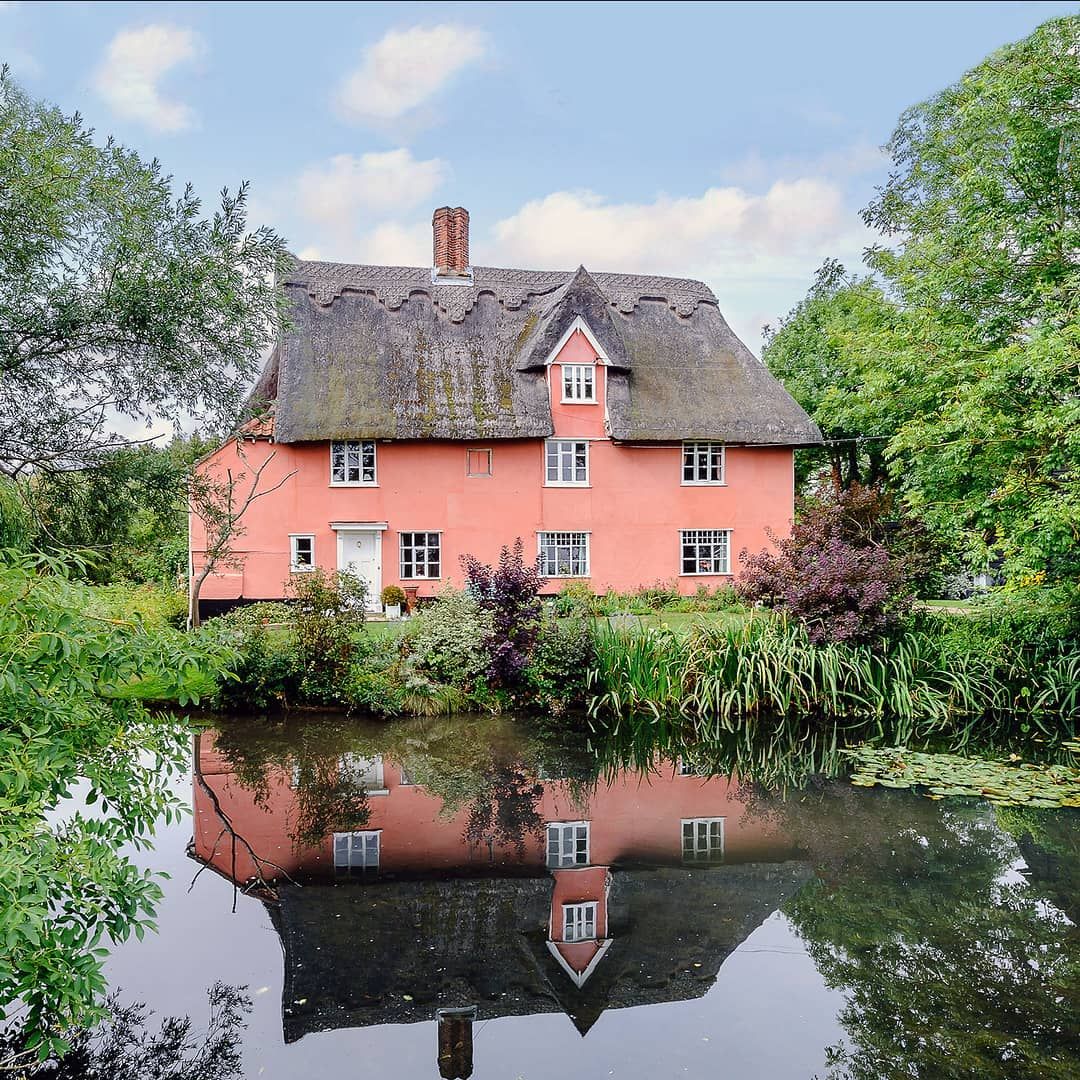 Pink thatched cottage beside still lake with mirror reflection, framed by trees and plants