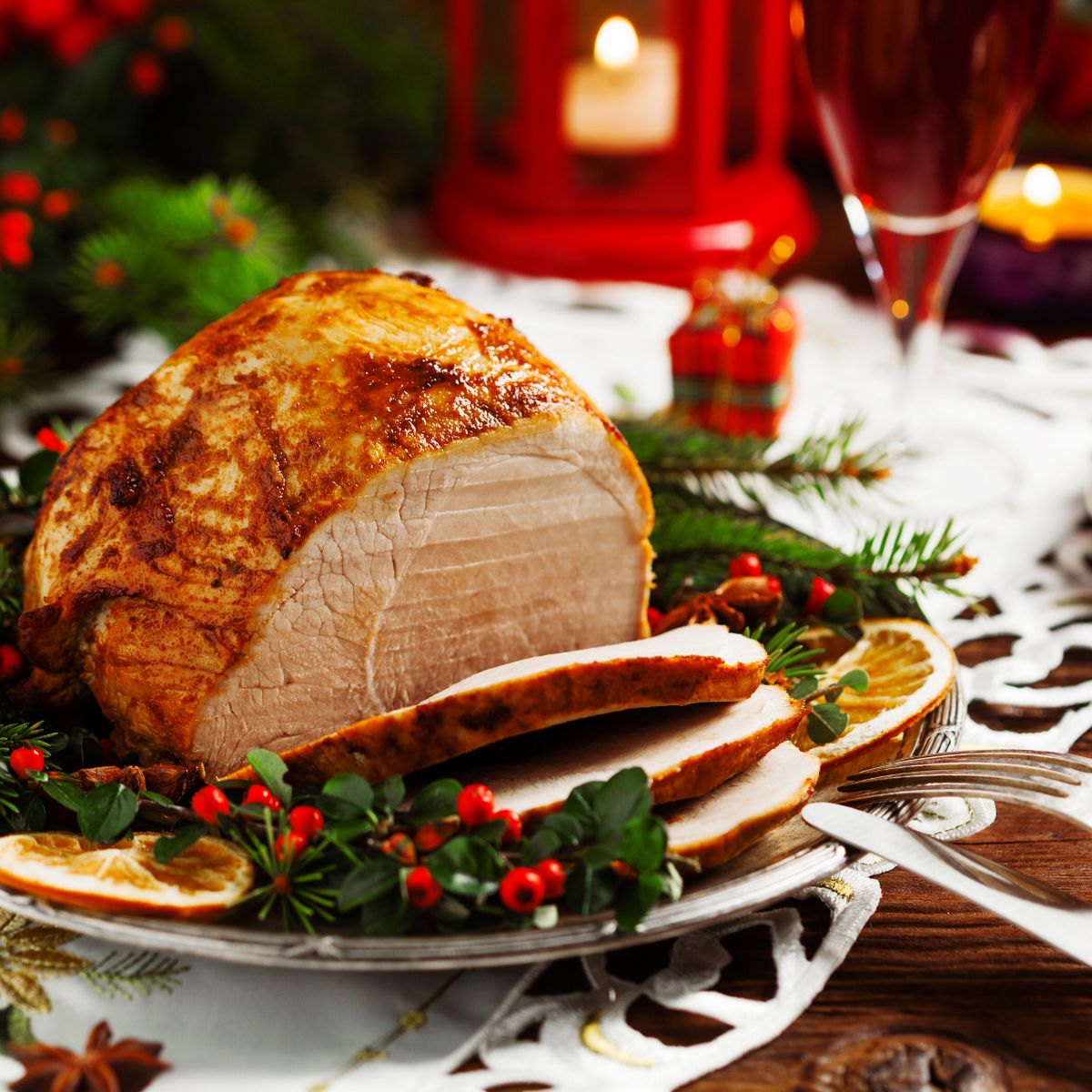 Sliced roast ham on a holiday table with holly, red berries, candle, and wine glass