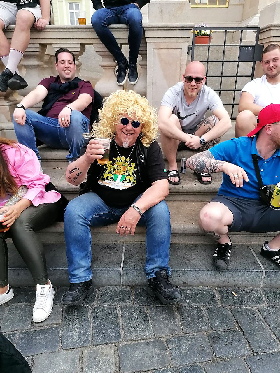 Man in blonde wig holding a drink sitting on stone steps with friends