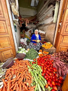 Woman selling fresh vegetables at a traditional market stall with carrots, tomatoes, and grens
