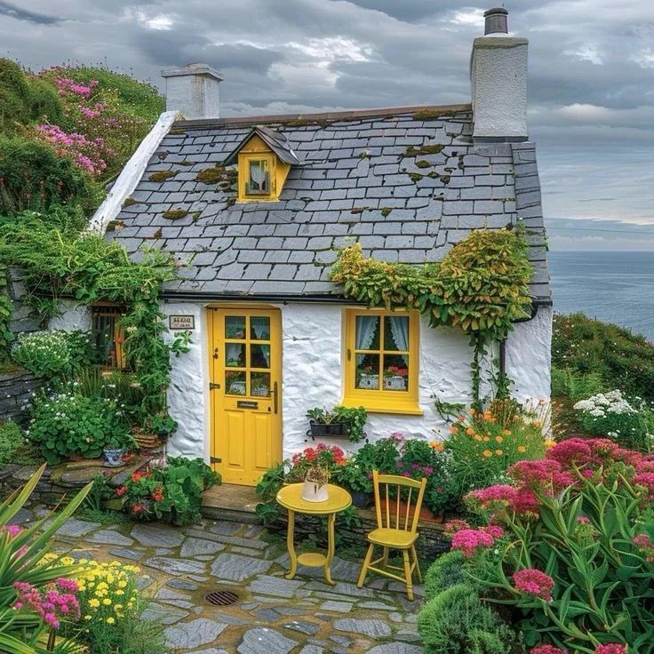 White seaside cottage with yellow door and windows, surrounded by colorful flowers and garden chairs