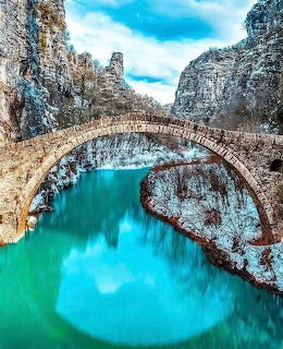 Old stone arch bridge over turquoise water in a snowy mountain gorge with tall cliffs