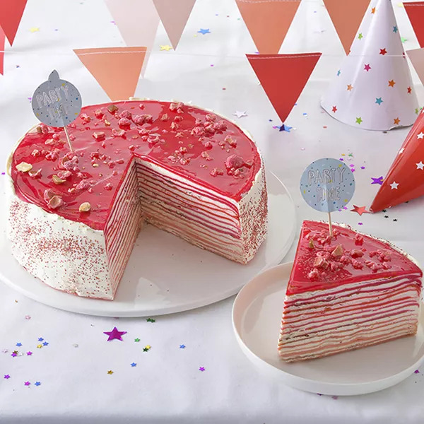Pink layered crepe cake on white plate with slice removed, party decorations in background