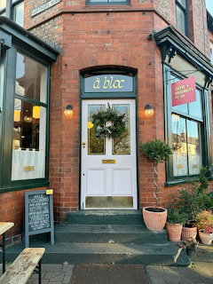 White front door of Café À Bloc with green wreath, red brick building, lanterns, and chalkboard sign