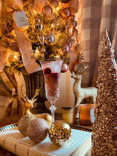 Gold Christmas ornaments, champagne flute, and decorated tree on rustic wooden table with warm lighting