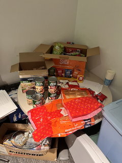 Clutered pantry with canned foods, snack boxes, chip bags, and supplies on a round table in a corner