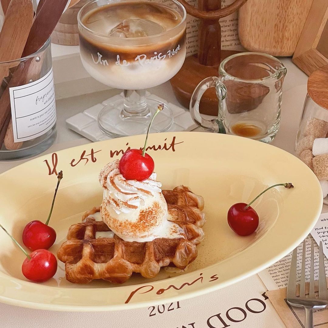 Waffle with whipped cream and cherry beside a glass of layered iced coffee on a table