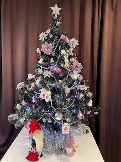 Christmas tree decorated with pink flowers, white ornaments, beaded garland, and star on top with figurines beneath