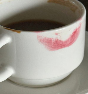 White coffee cup with dark coffee and a pink lipstick mark on the rim, sitting on a saucer.