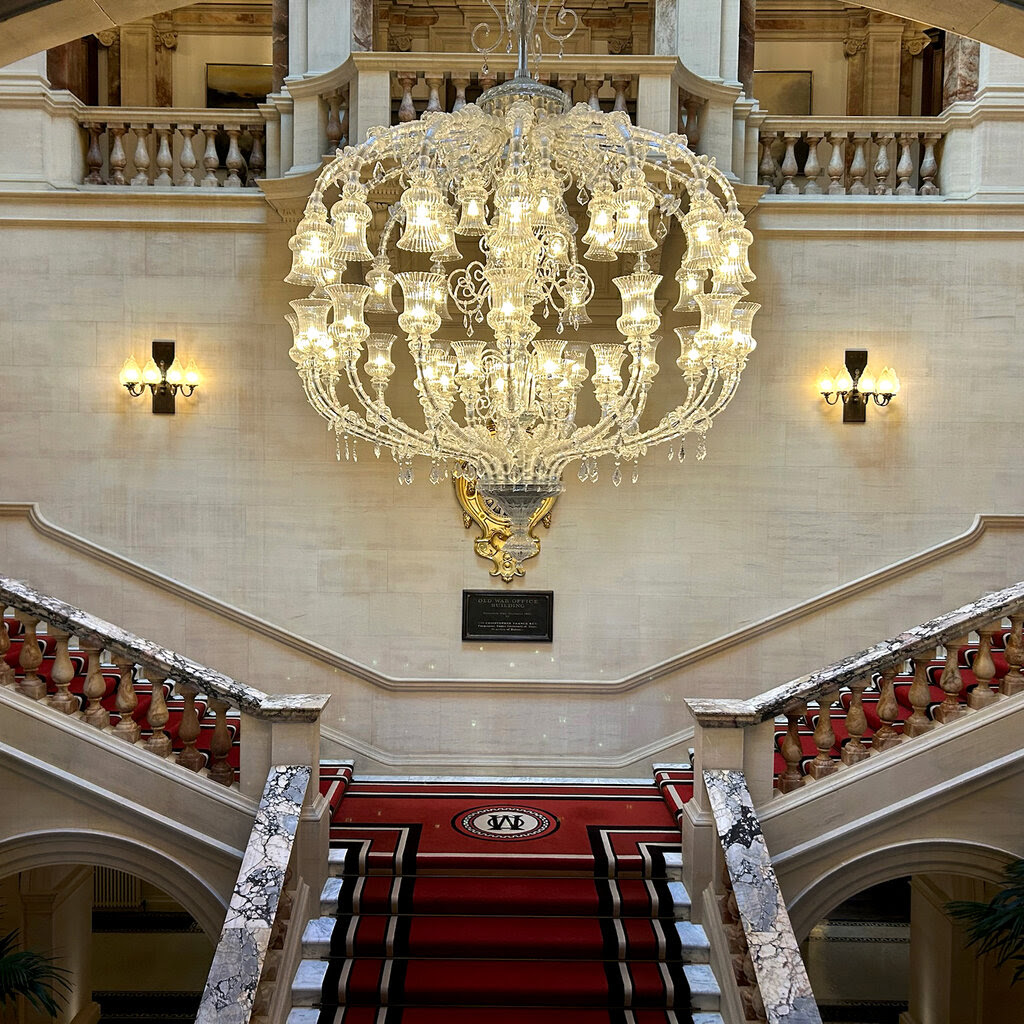 Ornate crystal chandelier above grand marble staircase with red carpet in elegant hotel lobby