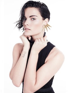 Dark-haired woman in black sleveless top posing with hand near face, wearing gold spike earrings