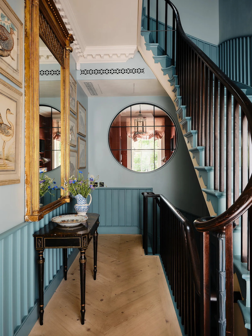 Elegant interior hallway with curved staircase, gold-framed mirror, round window, and wooden floor