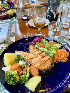 Grilled salmon fillet with vegetables and rice on blue plate, wine glasses on restaurant table