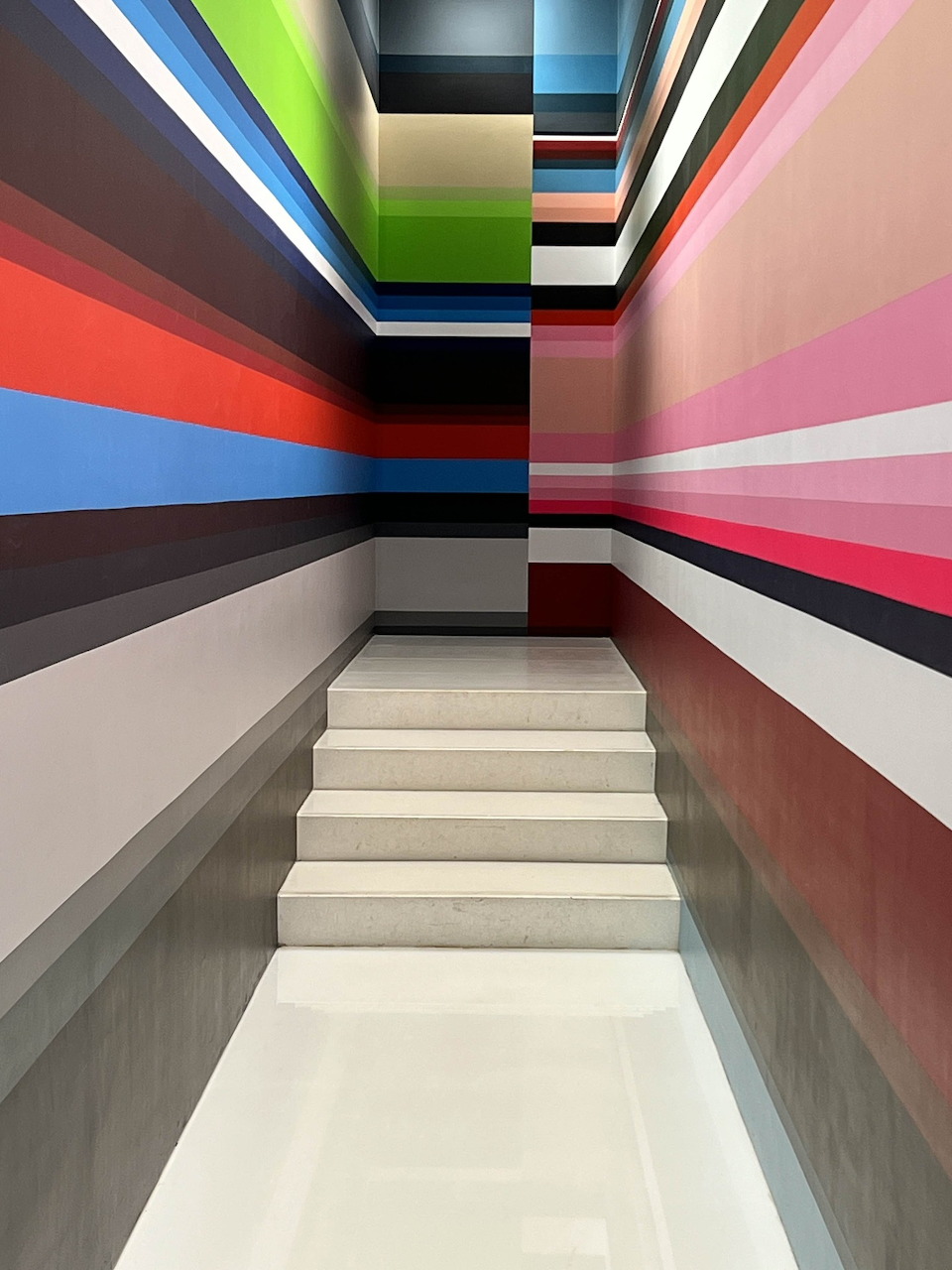 White stairs between walls with horizontal rainbow stripes in a narrow corridor