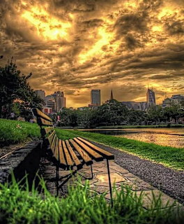 Wooden park bench along a riverside path at sunset with dramatic golden clouds and city skyline