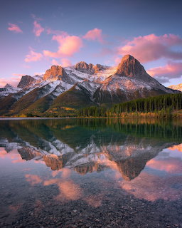 Snow-capped mountain peaks reflecting in a calm lake at sunset with pink clouds and evergreen trees along the shore