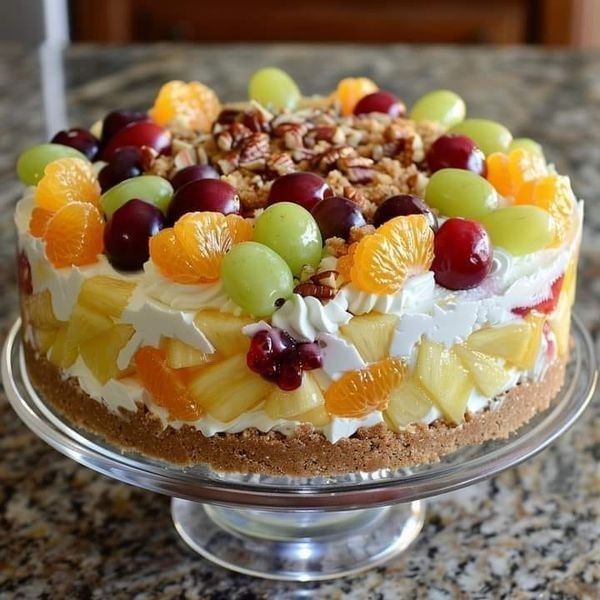 Round layered cake decorated with whipped cream, assorted fresh fruits, and nuts on a glass cake stand