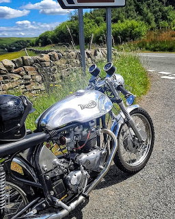 Silver Triton classic motorcycle on gravel roadside with stone wall and green countryside