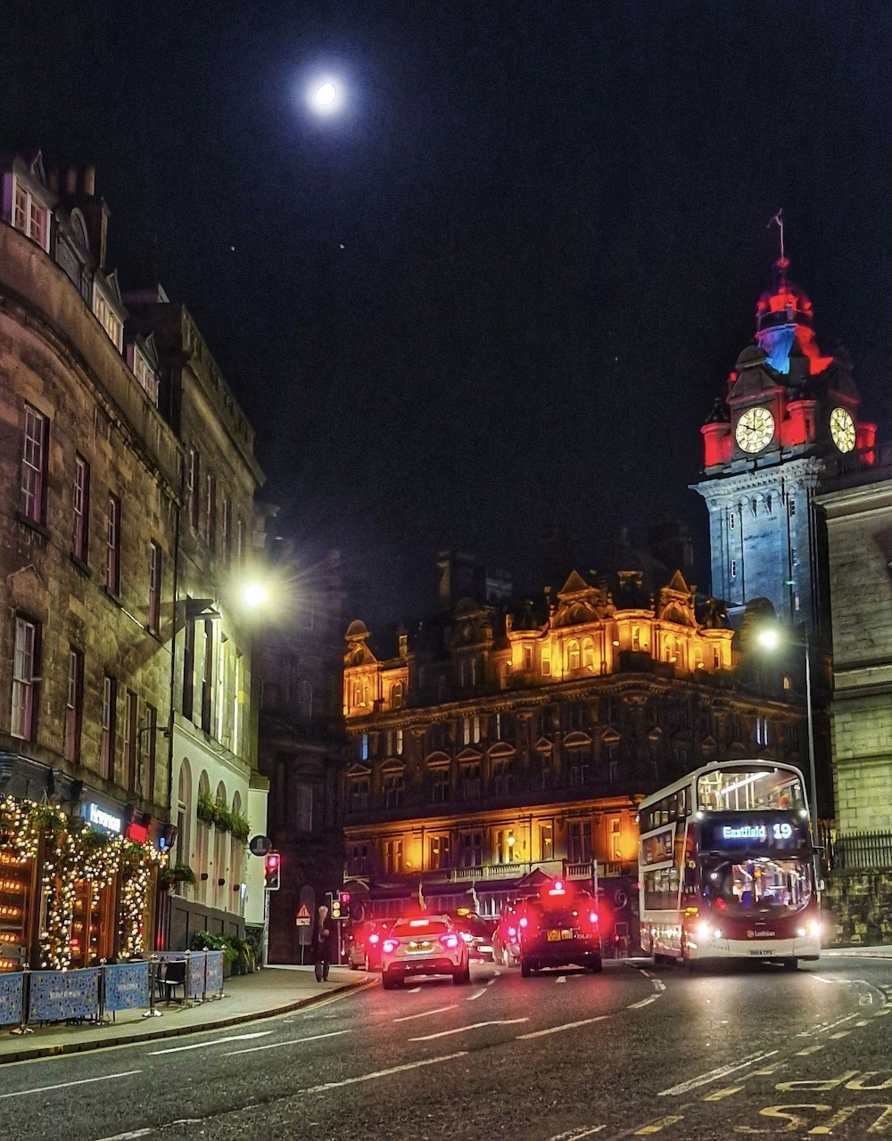 Edinburgh city street at night with lit castle on hill, decorated clock tower, and Christmas lights