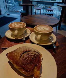 Two cappuccinos with latte art and a cinnamon roll pastry on a cafe table
