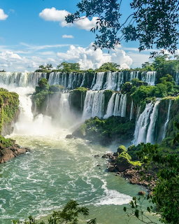 Multiple waterfalls flowing into green river surrounded by tropical vegetation and blue sky